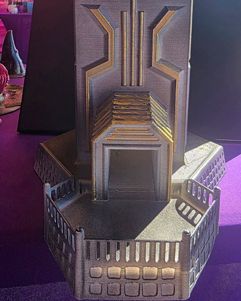 A tall gray dice tower with gold-painted trim, an arched exit gate, and a fenced base tray.