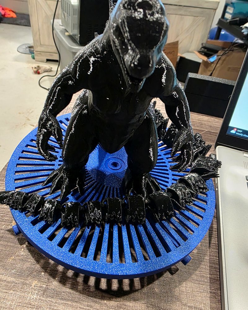 A large black 3D-printed Godzilla standing on a blue base, with a segmented tail curled around it.