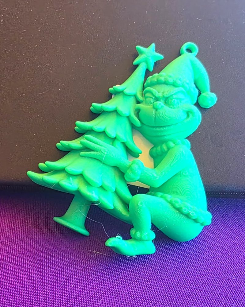 A green Grinch in a Santa hat hugging a Christmas tree, printed as a flat hanging ornament.