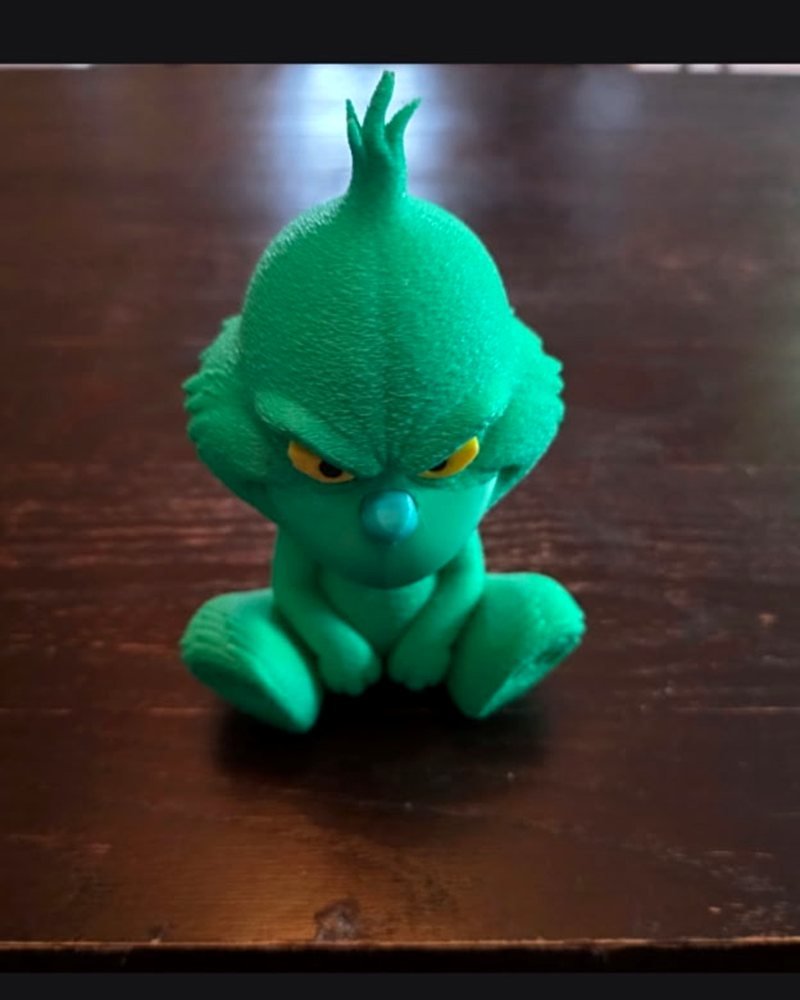 A small chibi-style Grinch figurine, printed in matte green PLA, sitting cross-legged on a wood table.