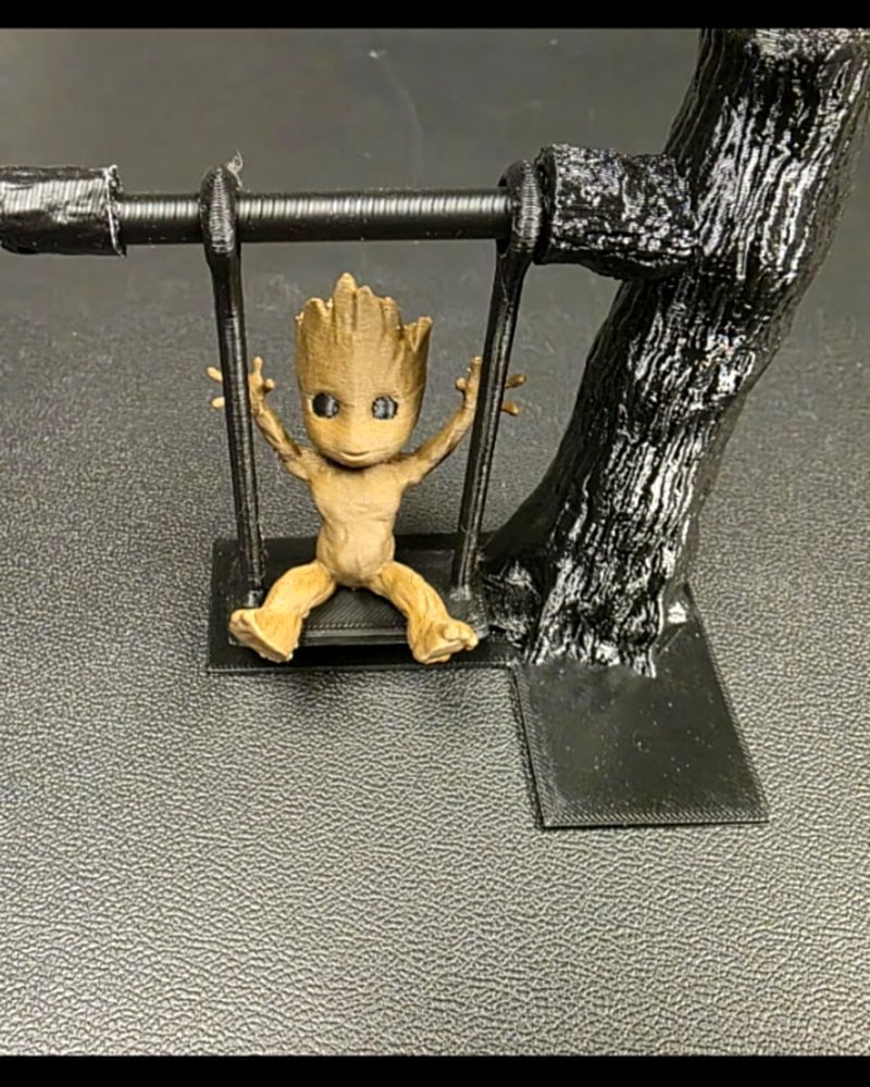 A small tan Groot figure sitting on a swing attached to a black tree trunk on a flat base.