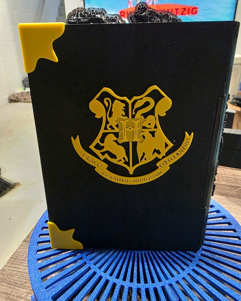 A black hardback-style book with a gold Hogwarts crest and gold corner caps, standing on a blue slatted plate.