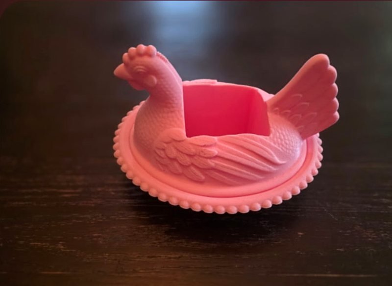 A pink hen-shaped bowl with a square hollow in its back, on a wood table.