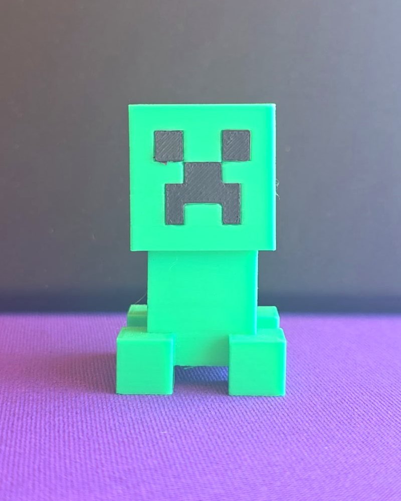 A small green Minecraft creeper figure with a black pixel face, on purple cloth.