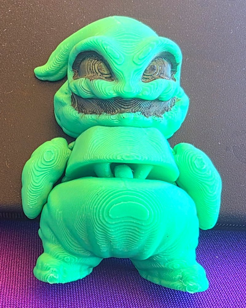 A small green Oogie Boogie figure with a toothy grin and hollow eyes, on purple cloth.