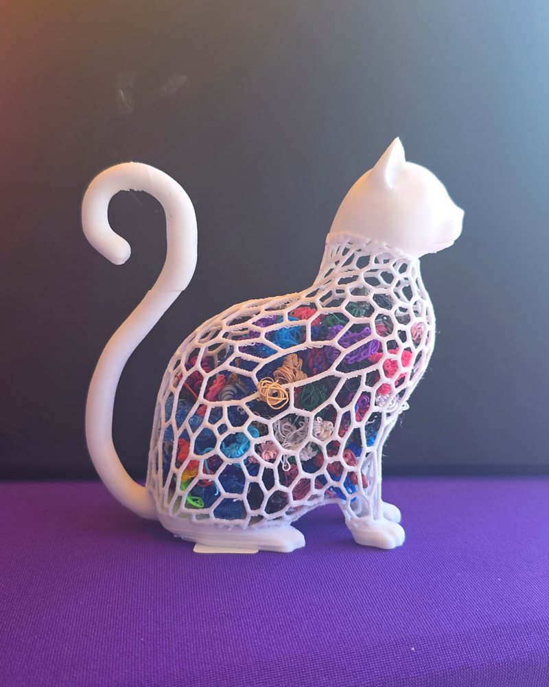 A white cat figurine with an openwork lattice body filled with colorful filament scraps, seen from the side.