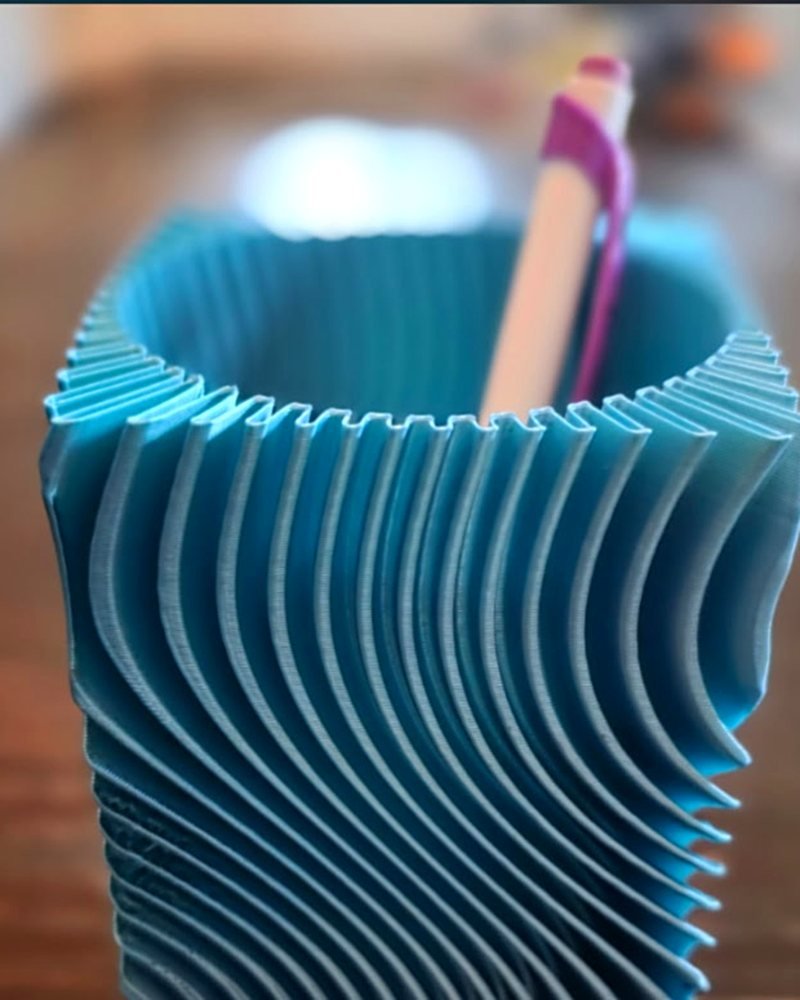A teal wavy-ribbed pen holder cup with a pen inside, on a desk.